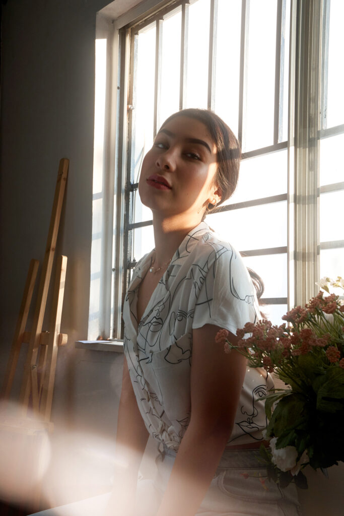 Nadya Inzunza a model with a contemplative expression sits by a window in a room with an artistic ambiance. She wears a white blouse with abstract line drawings and has a necklace with a pendant. The sunlight filters through the window bars casting a soft glow on her face.
